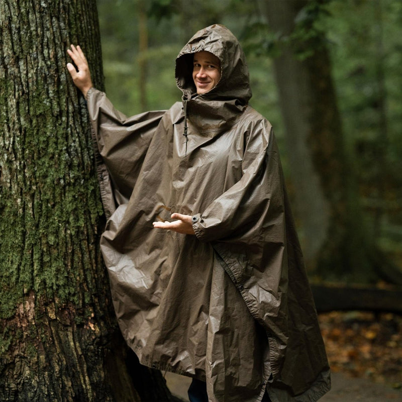 Man wearing vintage German army olive waterproof rain poncho with hood standing by tree in forest setting